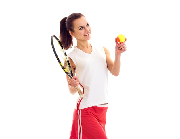 Joven mujer positiva con camisa deportiva blanca y falda roja sosteniendo raqueta de tenis y pelota amarilla