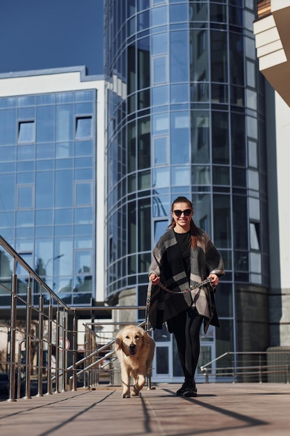 Joven mujer positiva se divierte con su perro cuando da un paseo al aire libre cerca del edificio de negocios