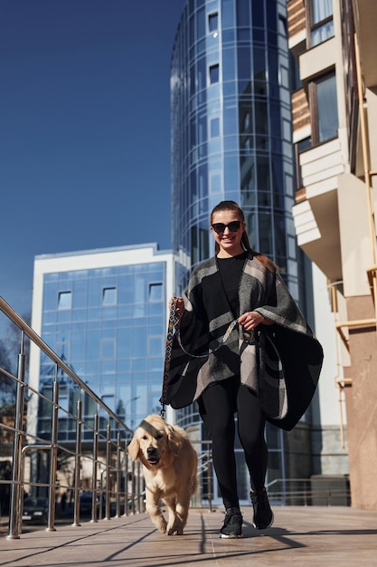 Joven mujer positiva se divierte con su perro cuando da un paseo al aire libre cerca del edificio de negocios