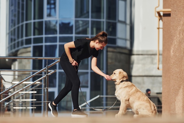 Foto joven mujer positiva se divierte con su perro cuando da un paseo al aire libre cerca del edificio de negocios