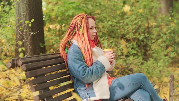 Joven mujer positiva con rastas de jengibre bebiendo café mirando hacia otro lado sentada en un banco del parque en otoño dorado