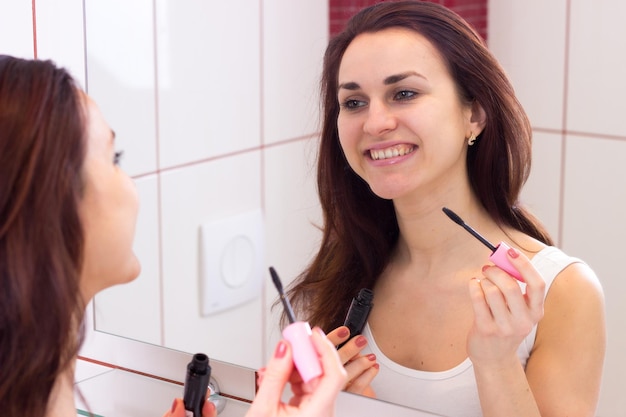 Joven mujer positiva usando rímel rosa frente al espejo en su baño burdeos