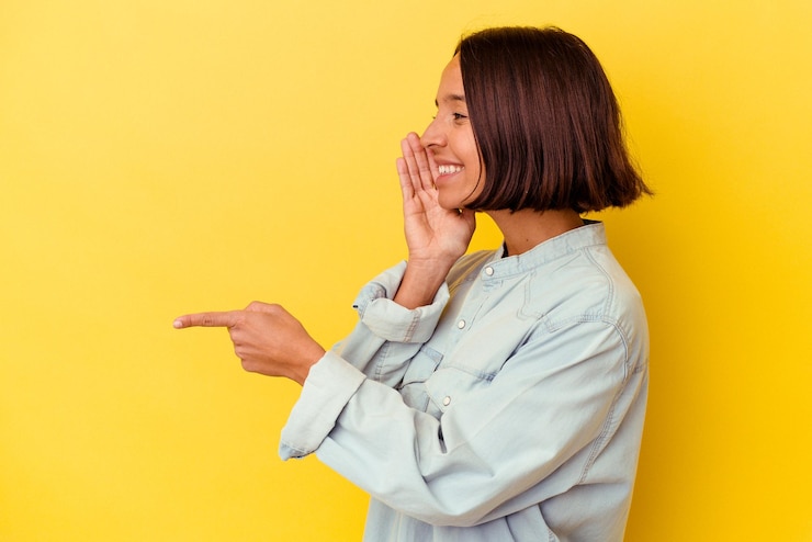 Foto joven mujer de raza mixta aislada de fondo amarillo diciendo un chisme, señalando al lado informando algo.