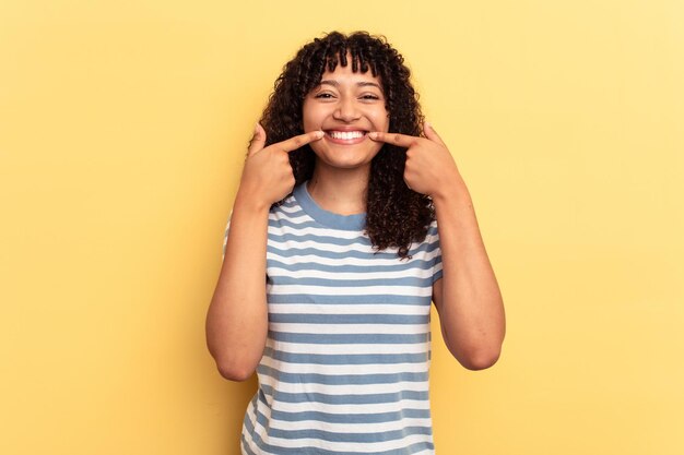 Joven mujer de raza mixta aislada de fondo amarillo sonríe señalando con el dedo a la boca