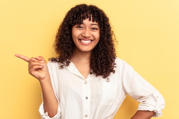 Joven mujer de raza mixta aislada de fondo amarillo sonriendo alegremente señalando con el dedo índice lejos