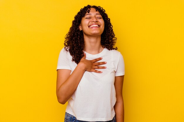 Joven mujer de raza mixta aislada sobre fondo amarillo se ríe a carcajadas manteniendo la mano en el pecho.