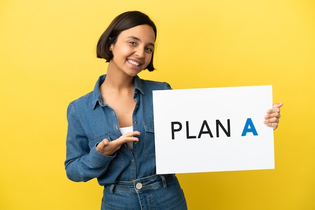 Joven mujer de raza mixta aislada sobre fondo amarillo sosteniendo un cartel con el mensaje PLAN A y apuntando