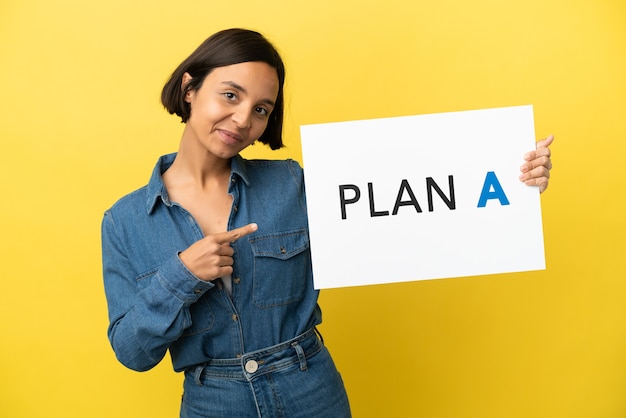 Joven mujer de raza mixta aislada sobre fondo amarillo sosteniendo un cartel con el mensaje PLAN A y apuntando