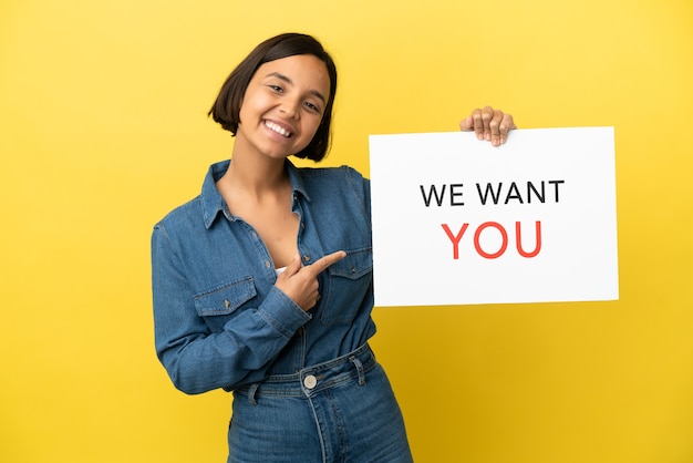 Joven mujer de raza mixta aislada sobre fondo amarillo sosteniendo We Want You board y apuntando