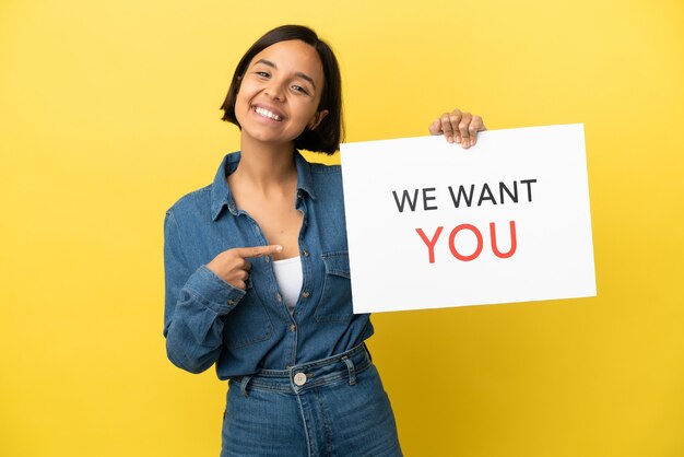 Joven mujer de raza mixta aislada sobre fondo amarillo sosteniendo We Want You board y apuntando