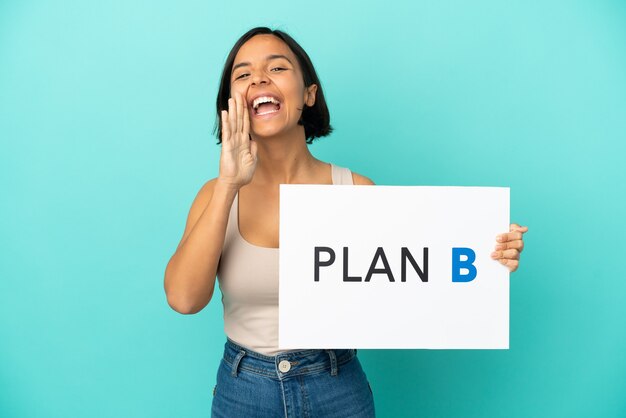 Joven mujer de raza mixta aislada sobre fondo azul sosteniendo un cartel con el mensaje PLAN B y gritando