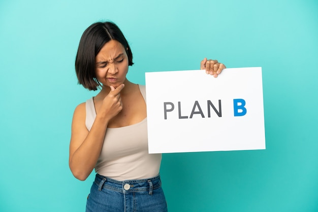 Joven mujer de raza mixta aislada sobre fondo azul sosteniendo un cartel con el mensaje PLAN B y pensando