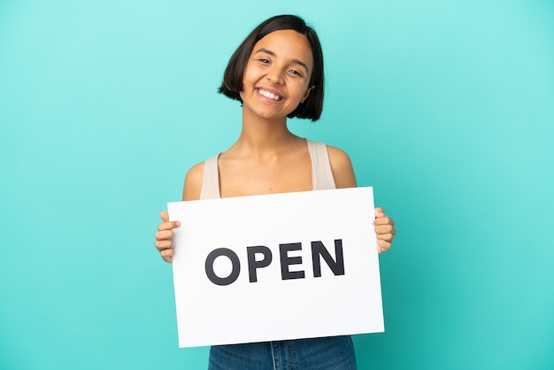 Joven mujer de raza mixta aislada sobre fondo azul sosteniendo un cartel con texto ABIERTO con expresión feliz