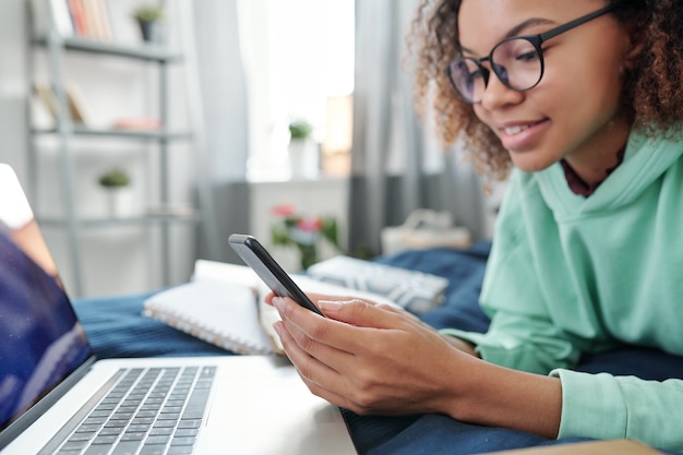 Foto joven mujer de raza mixta en anteojos desplazándose en el teléfono inteligente mientras está acostado en la cama frente a la computadora portátil y preparando la tarea en casa