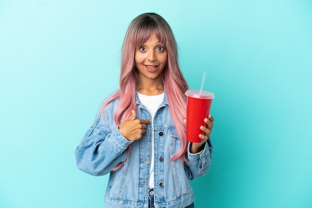 Joven mujer de raza mixta bebiendo una bebida fresca aislada sobre fondo azul con expresión facial sorpresa