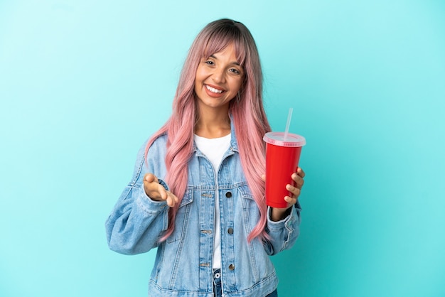 Joven mujer de raza mixta bebiendo una bebida fresca aislado sobre fondo azul un apretón de manos para cerrar un buen trato