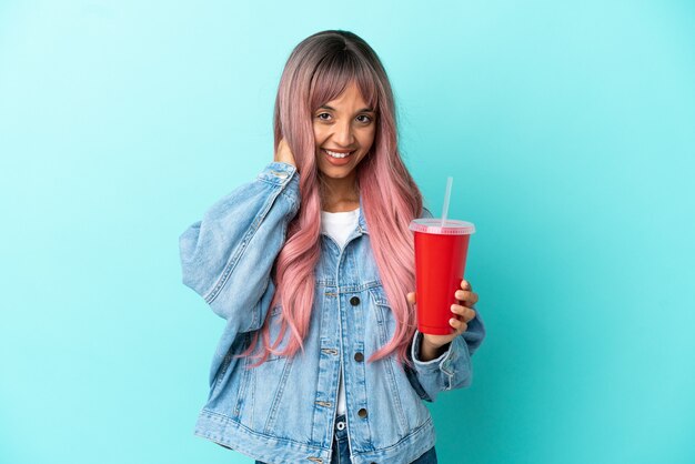 Joven mujer de raza mixta bebiendo una bebida fresca aislado sobre fondo azul riendo