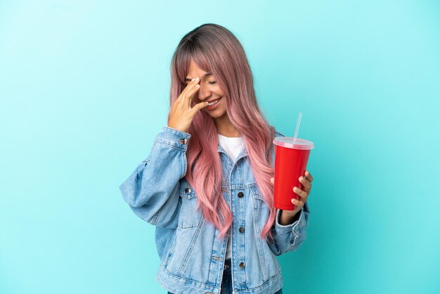 Joven mujer de raza mixta bebiendo una bebida fresca aislado sobre fondo azul riendo