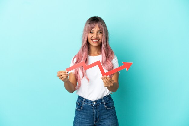 Joven mujer de raza mixta con cabello rosado aislado sobre fondo azul sosteniendo una captura de una flecha ascendente con expresión feliz