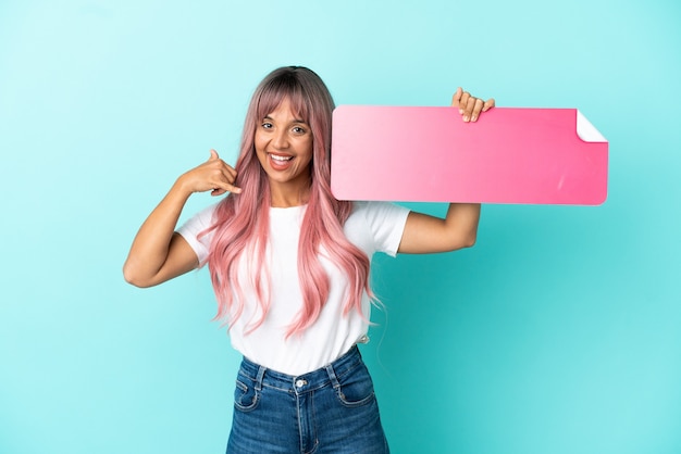 Joven mujer de raza mixta con cabello rosado aislado sobre fondo azul sosteniendo un cartel vacío y haciendo gesto de teléfono