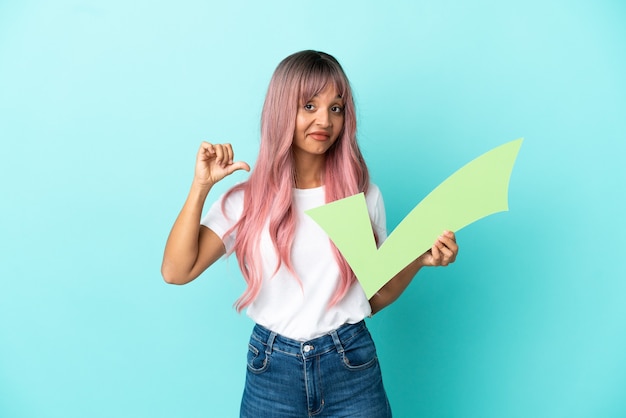 Joven mujer de raza mixta con cabello rosado aislado sobre fondo azul sosteniendo un icono de verificación con gesto orgulloso