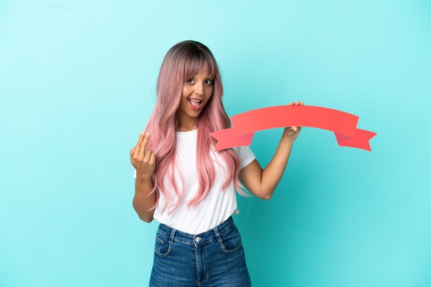 Joven mujer de raza mixta con cabello rosado aislado sobre fondo azul sosteniendo una pancarta vacía y haciendo el gesto que viene