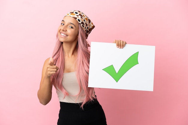 Joven mujer de raza mixta con cabello rosado aislado sobre fondo rosa sosteniendo un cartel con el icono de marca de verificación verde de texto y apuntando hacia el frente