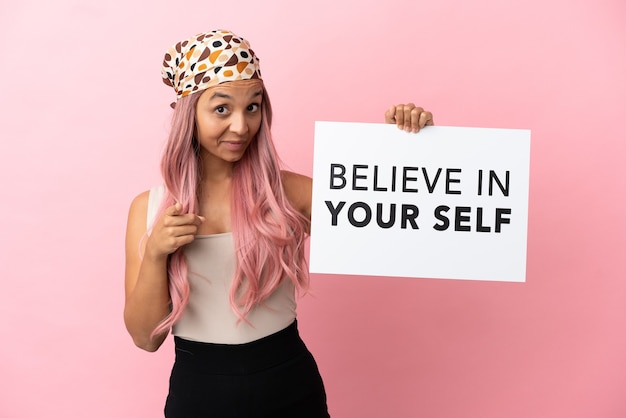 Foto joven mujer de raza mixta con cabello rosado aislado sobre fondo rosa sosteniendo un cartel con el texto cree en ti mismo y apuntando hacia el frente