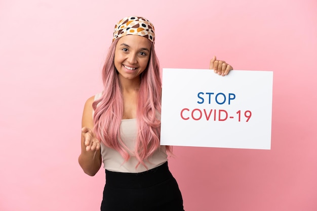 Joven mujer de raza mixta con cabello rosado aislado sobre fondo rosa sosteniendo un cartel con texto Stop Covid 19 haciendo un trato