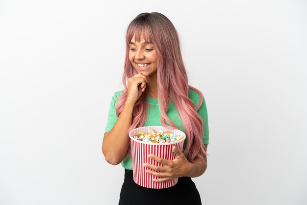 Joven mujer de raza mixta con cabello rosado comiendo palomitas de maíz aislado sobre fondo blanco mirando hacia el lado y sonriendo