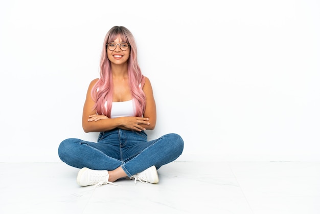 Joven mujer de raza mixta con cabello rosado sentada en el suelo aislado sobre fondo blanco con los brazos cruzados y mirando hacia adelante