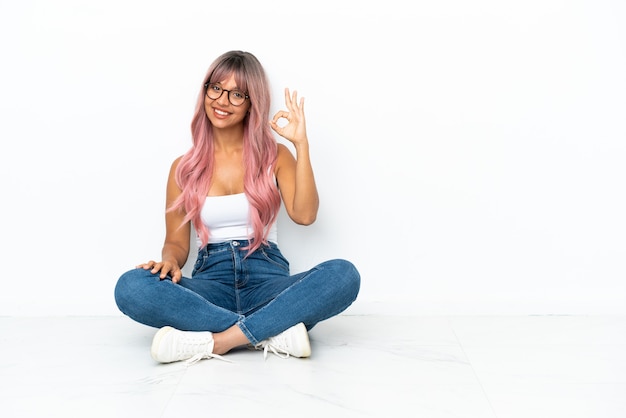 Joven mujer de raza mixta con cabello rosado sentada en el suelo aislado sobre fondo blanco mostrando el signo de ok con los dedos