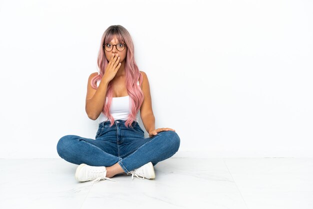 Joven mujer de raza mixta con cabello rosado sentada en el suelo aislado sobre fondo blanco sorprendida y conmocionada mientras mira a la derecha