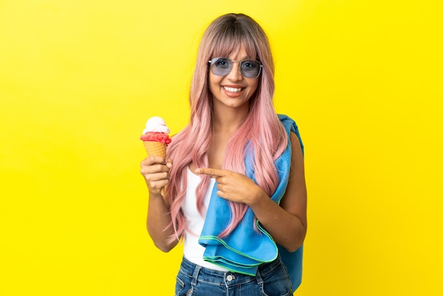 Joven mujer de raza mixta con cabello rosado sosteniendo helado aislado sobre fondo amarillo apuntando hacia el lado para presentar un producto