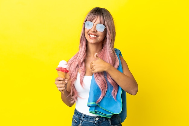 Joven mujer de raza mixta con cabello rosado sosteniendo helado aislado sobre fondo amarillo dando un pulgar hacia arriba gesto