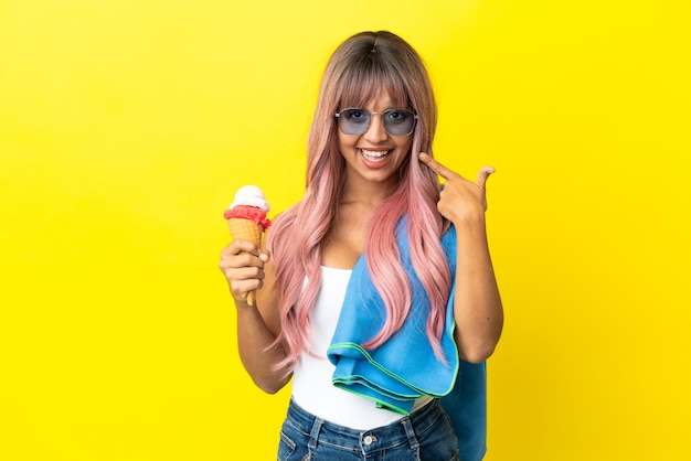 Joven mujer de raza mixta con cabello rosado sosteniendo helado aislado sobre fondo amarillo dando un pulgar hacia arriba gesto