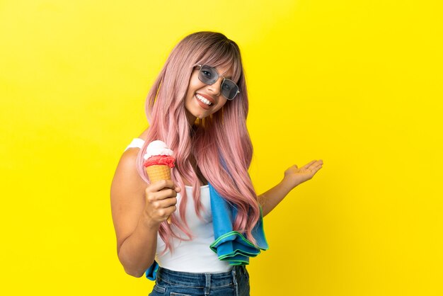 Joven mujer de raza mixta con cabello rosado sosteniendo helado aislado sobre fondo amarillo extendiendo las manos hacia el lado para invitar a venir