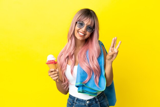 Joven mujer de raza mixta con cabello rosado sosteniendo helado aislado sobre fondo amarillo feliz y contando tres con los dedos