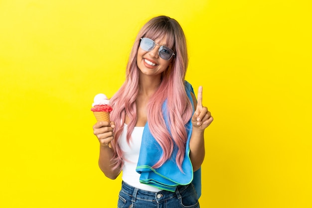 Joven mujer de raza mixta con cabello rosado sosteniendo helado aislado sobre fondo amarillo mostrando y levantando un dedo