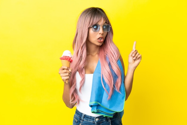 Joven mujer de raza mixta con cabello rosado sosteniendo helado aislado sobre fondo amarillo pensando en una idea apuntando con el dedo hacia arriba