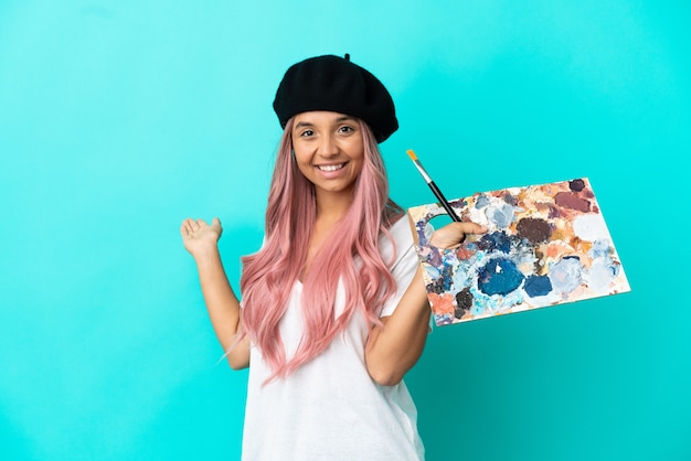 Foto joven mujer de raza mixta con cabello rosado sosteniendo una paleta aislada sobre fondo azul extendiendo las manos hacia el lado para invitar a venir