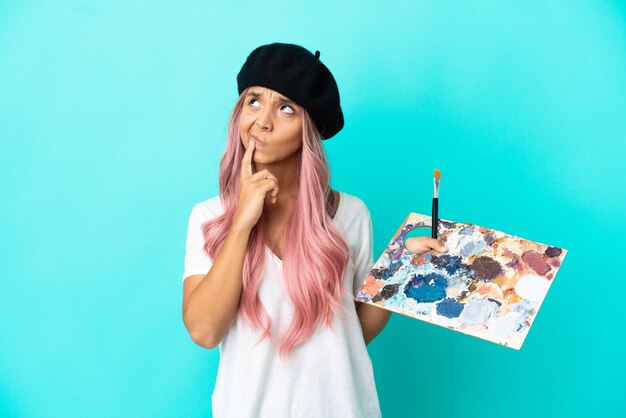 Joven mujer de raza mixta con cabello rosado sosteniendo una paleta aislada sobre fondo azul teniendo dudas mientras mira hacia arriba