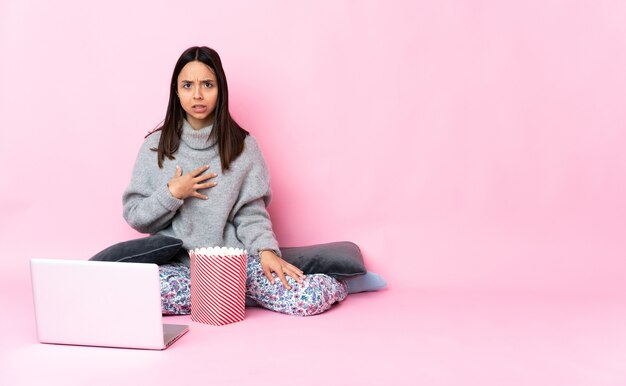 Joven mujer de raza mixta comiendo palomitas de maíz mientras ve una película en la computadora portátil apuntando a sí mismo