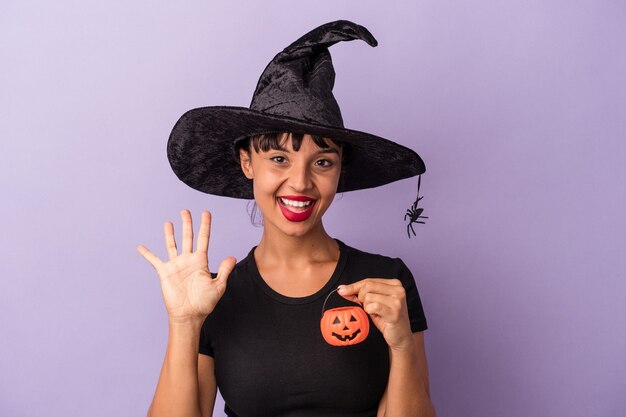 Joven mujer de raza mixta disfrazada de bruja aislada sobre fondo púrpura sonriendo alegre mostrando el número cinco con los dedos.