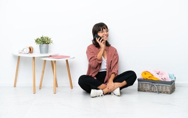 Joven mujer de raza mixta doblando ropa sentada en el suelo aislada de fondo blanco manteniendo una conversación con el teléfono móvil