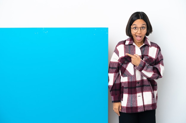 Foto joven mujer de raza mixta con un gran cartel azul aislado sobre fondo blanco sorprendido y apuntando hacia el lado