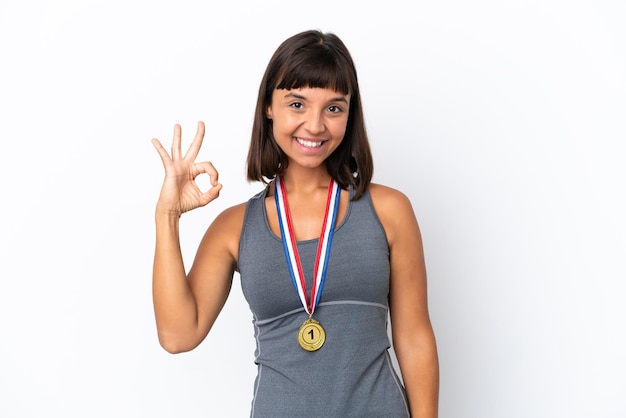 Joven mujer de raza mixta con medallas aislado sobre fondo blanco mostrando signo ok con los dedos