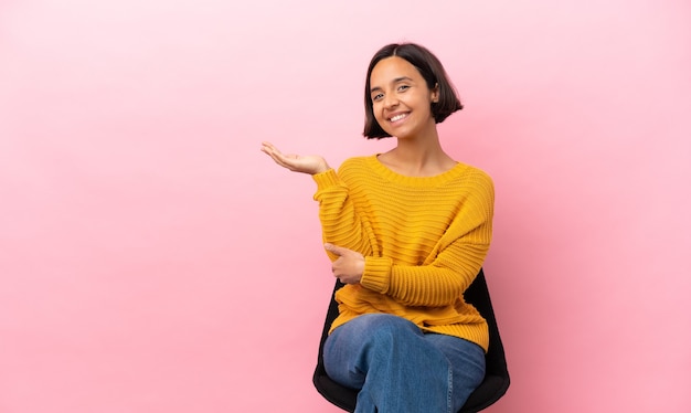 Joven mujer de raza mixta sentada en una silla aislada extendiendo las manos hacia el lado para invitar a venir