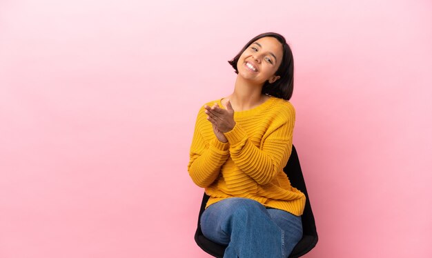 Joven mujer de raza mixta sentada en una silla aislada sobre fondo rosa aplaudiendo