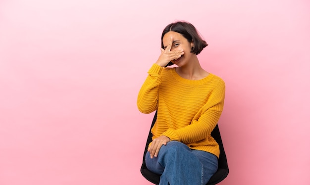 Joven mujer de raza mixta sentada en una silla aislada sobre fondo rosa cubriéndose los ojos con las manos y sonriendo